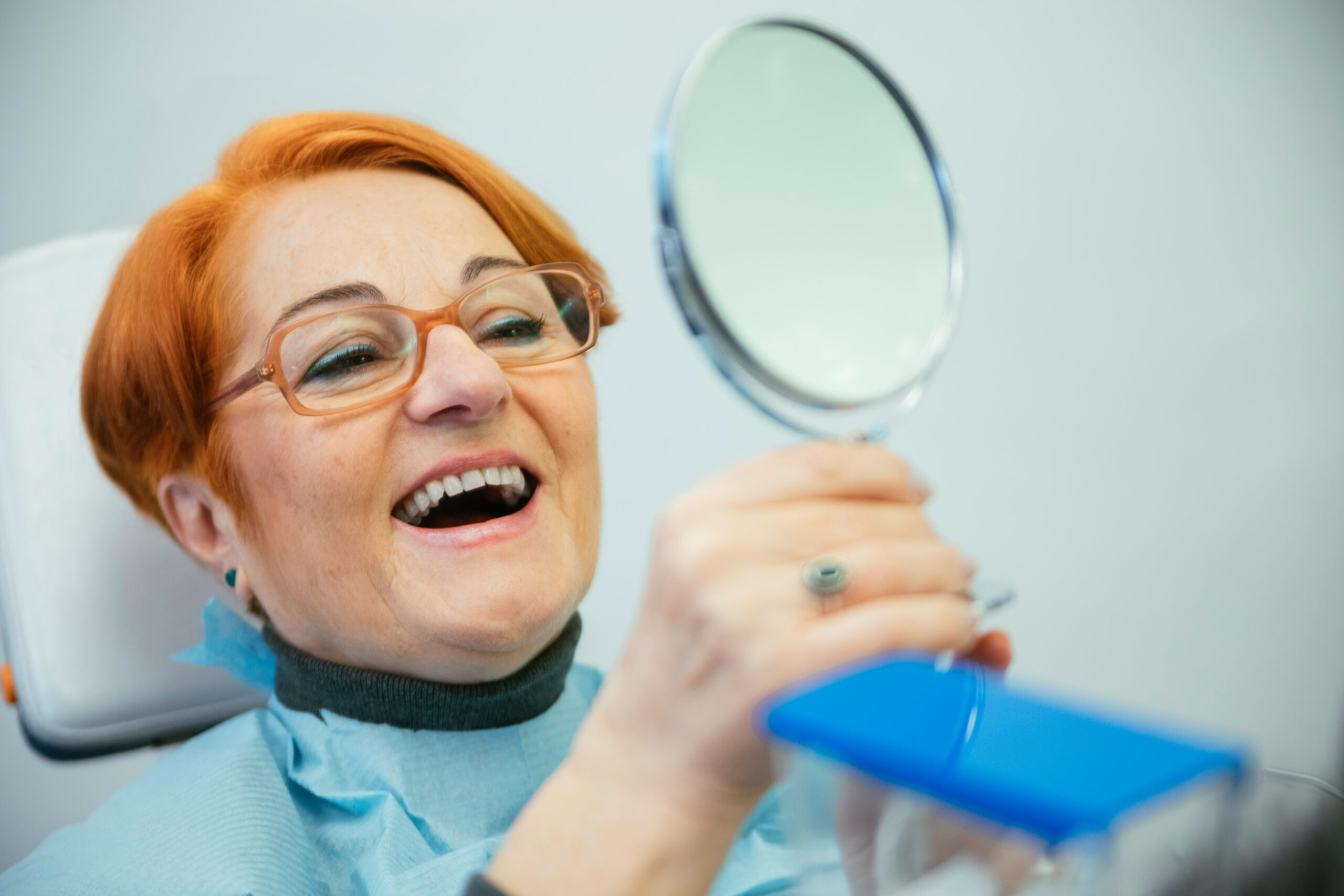 Senior Woman Sitting In A Chair Looking At Her Teeth with partial dentures In The Mirror